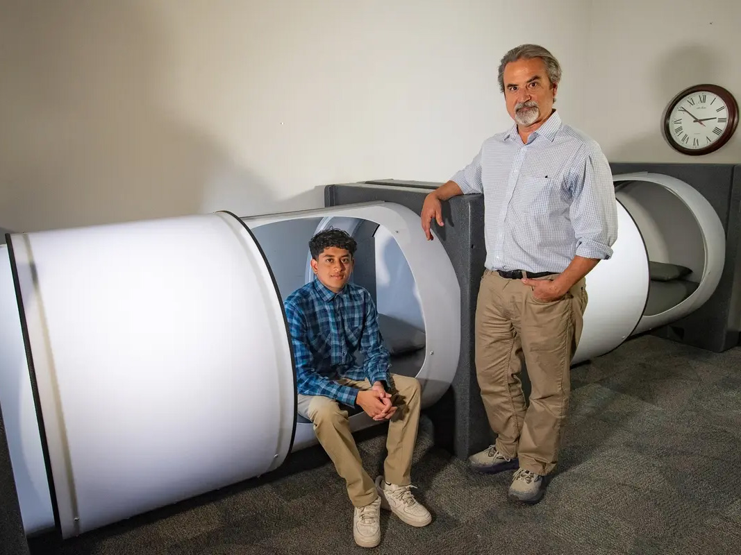 A man sitting in a white pod, and another man standing to the right of the pod, resting his left arm on the side of it.