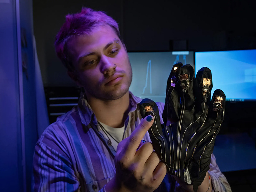A student wearing a black glove with metallic wires on it, on his right hand. His left index finger is touching his right thumb.