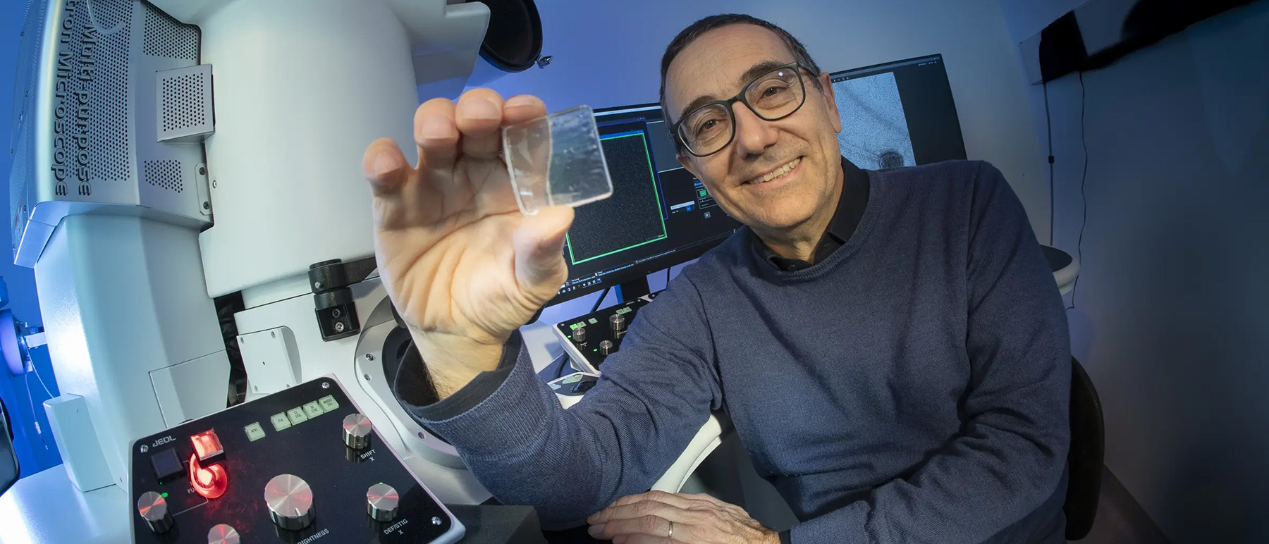 Physics professor Massimo Bertino holds up a small piece of glass in a room with computers and scientific equipment