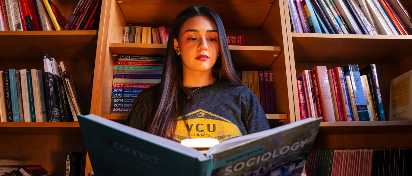 A woman reads an open book in front of a bookshelf