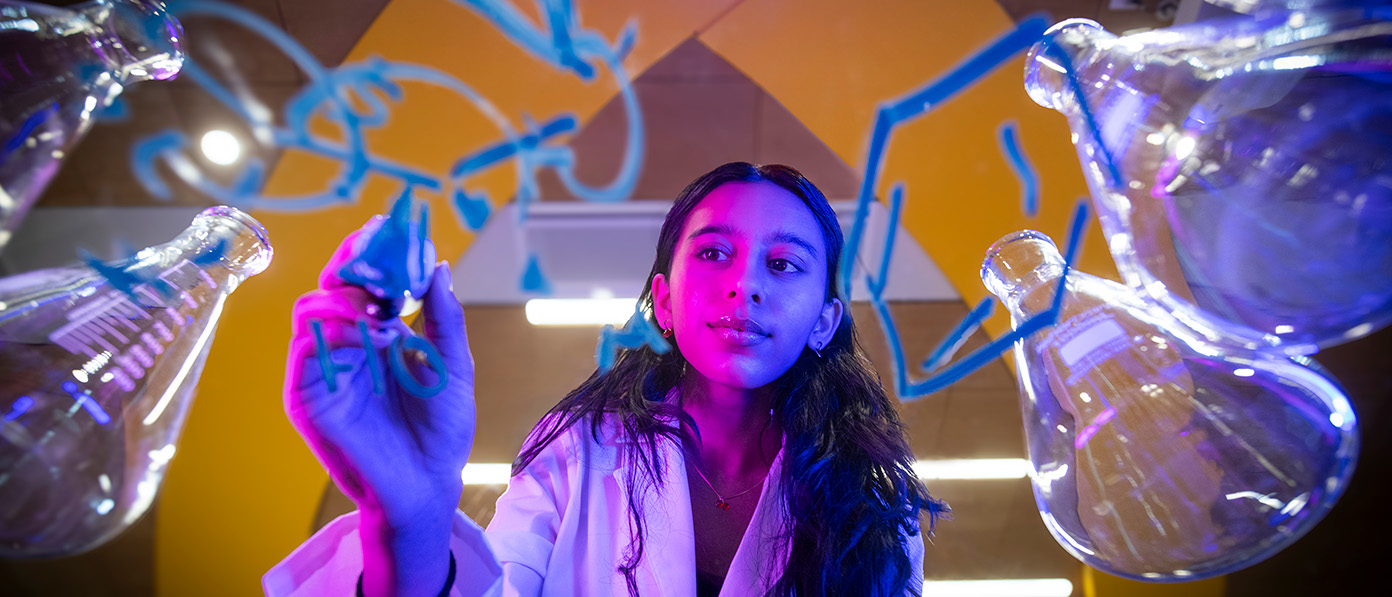a student writes an equation on a glass table surrounded by beakers