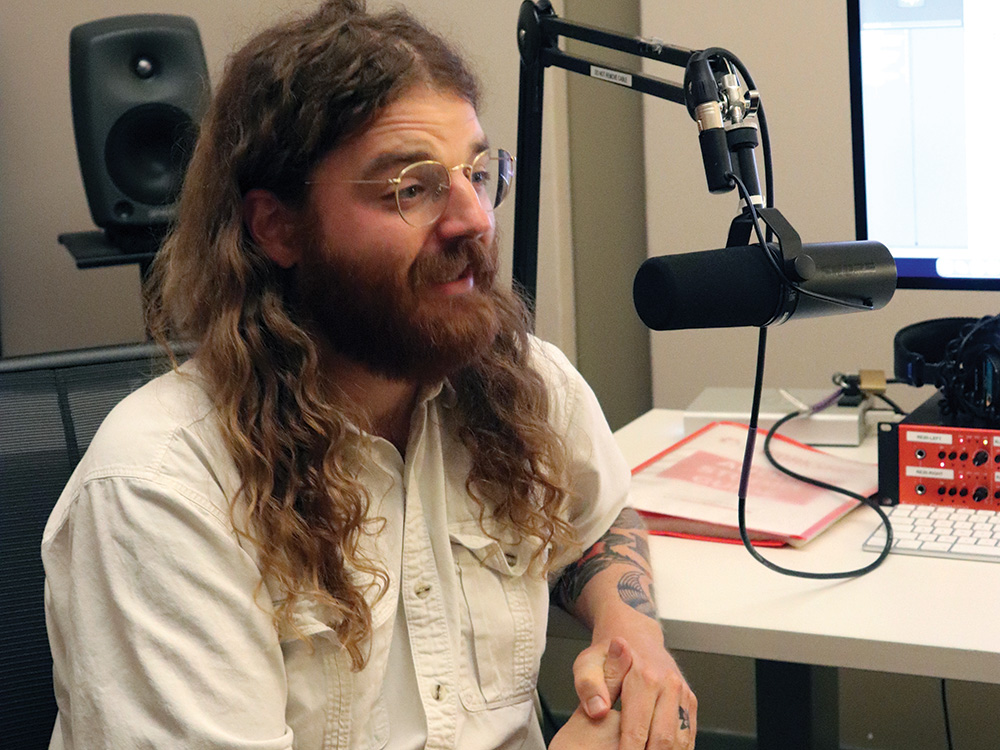 Lead editor of Blackbird, Trey Burnart Hall, sits at a desk speaking into a microphone