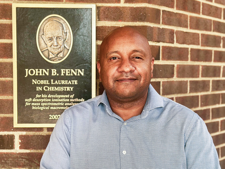 Fabio Gomes stands in front of a brick wall with a plaque of John Fenn