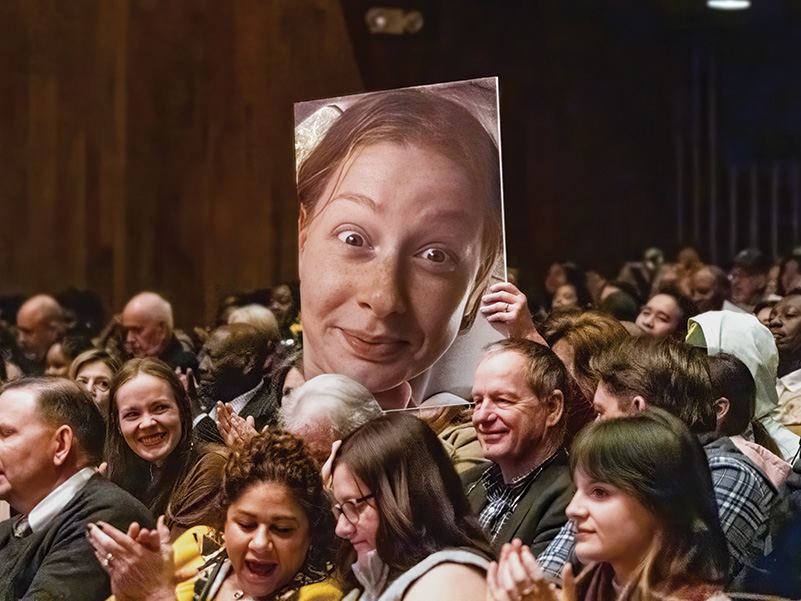 An audience member in an auditorium holds up a large photo of a woman's face