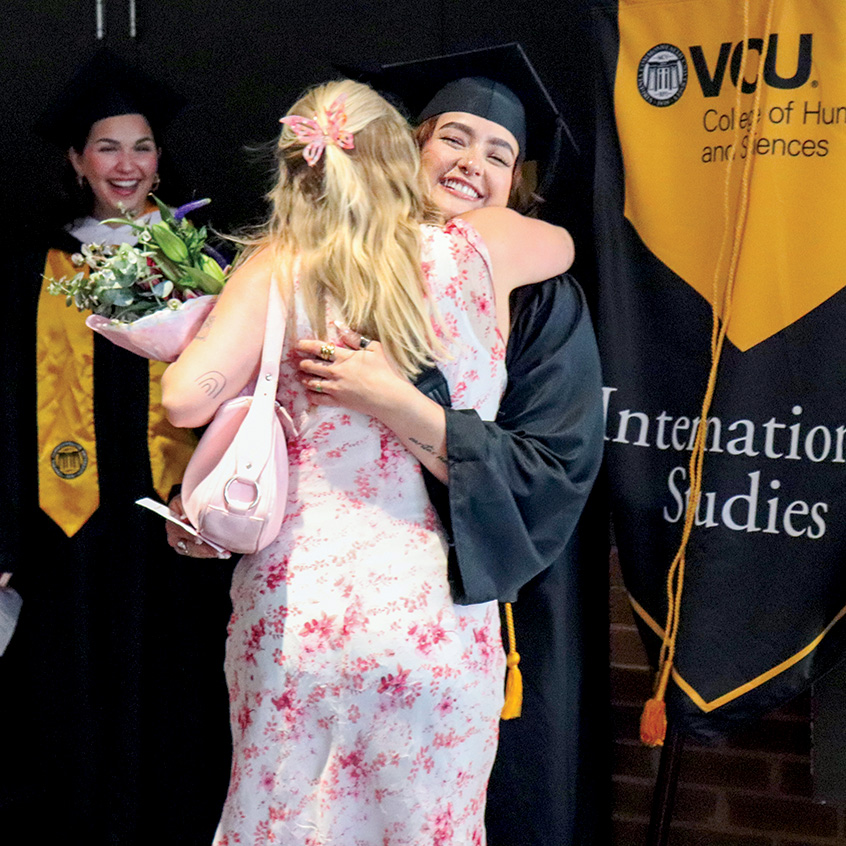 A student in graduation regalia embraces a woman in a dress in front of a VCU International Studies banner while another graduate looks on smiling