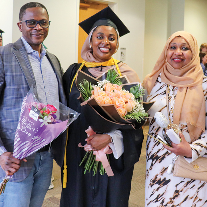 A student in graduation regalia holding a bouquet of flowers stands between a man in a suit and a woman in a dress and head scarf
