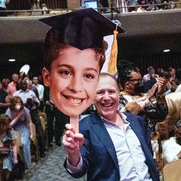 Man sitting in an audience holding a cutout head of a boy wearing a graduation cap