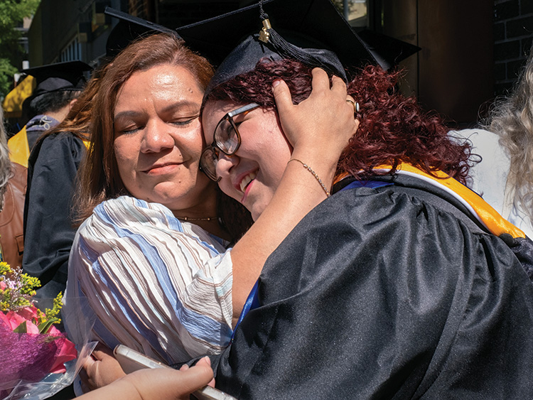 A woman in graduation regalia hugs another woman