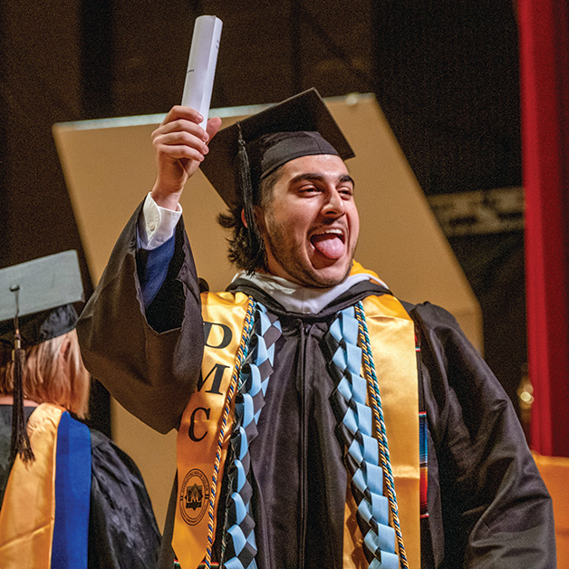 A student in graduation regalia holds up his diploma and sticks out his tongue