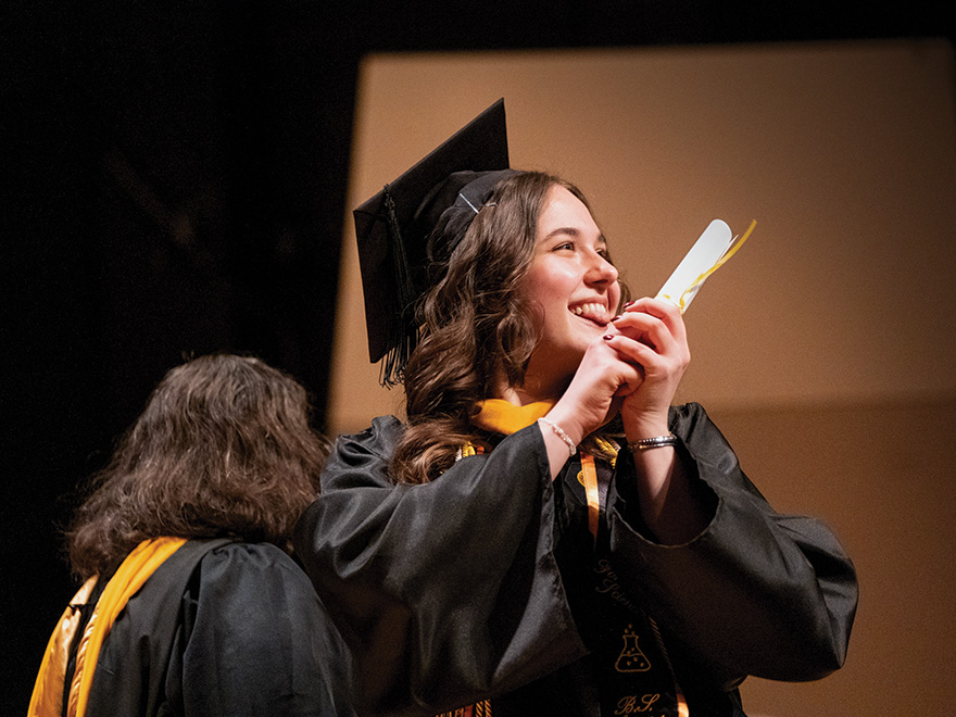 A woman in graduation regalia smiles and holds a diploma