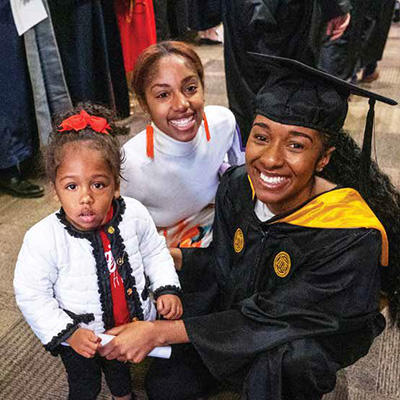 A student in graduation regalia kneeling beside a woman and young girl