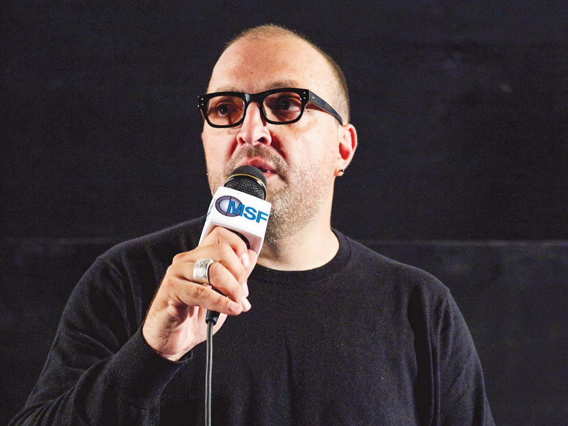 Rob Milazzo speaks into a microphone against a dark backdrop