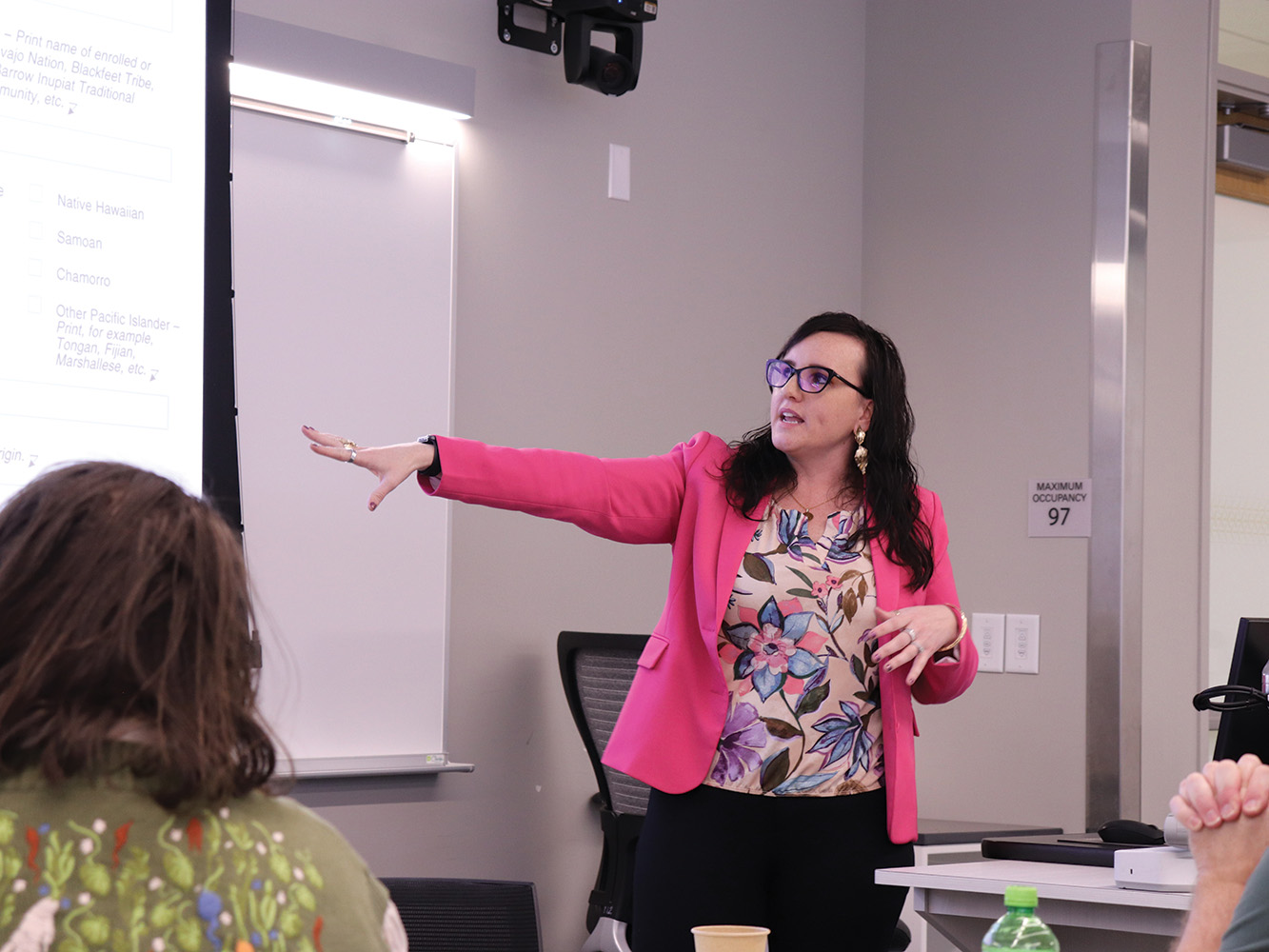 A woman in a pink blazer points to a projector screen in a classroom
