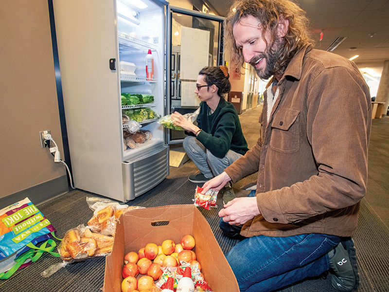 A man knees on the floor in front of a crate of apples; behind him, a woman places food inside a fridge