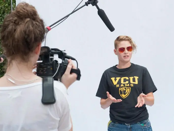 A photo of a woman speaking while standing in front of a white background. In front of her, a man with a microphone holds it above her head, a woman points a video camera at her and a man stands to the side.