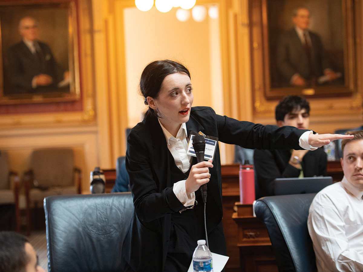 A woman in a black sport coat stands in a Virginia Capitol chambers room, speaking a microphone and extends her arm
