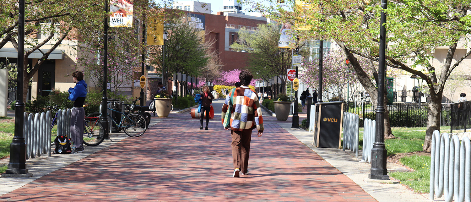 A person in a multicolored sweater walks down a brick sidewalk on VCU's campus, facing away from the camera