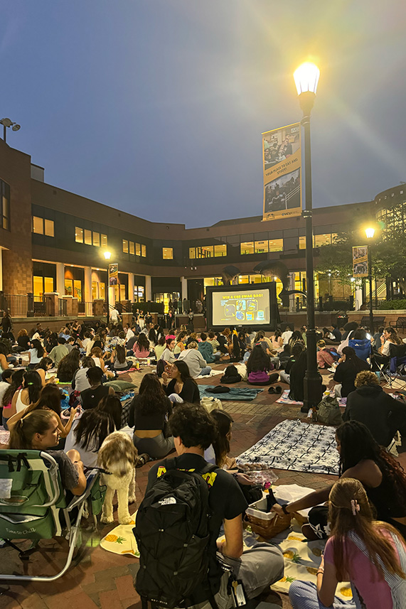 A large group of students sit in the VCU Commons courtyard in front of a large projector screen at dusk