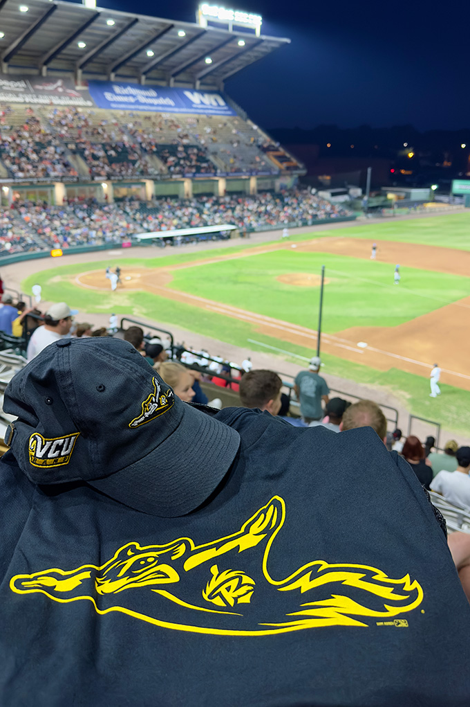 A black and gold Richmond Flying Squirrels hat and t-shirt sit in the foreground with a baseball diamond, stadium seating and the night sky in the background