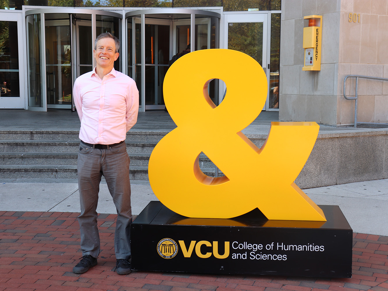 Peter Henry stands beside a gold ampersand statue in front of Cabell Library