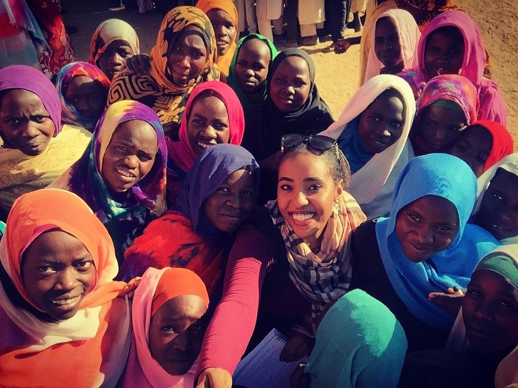 Isra'a Nour surrounded by a large group of smiling children and women wearing vibrant, colorful hijabs and headscarves